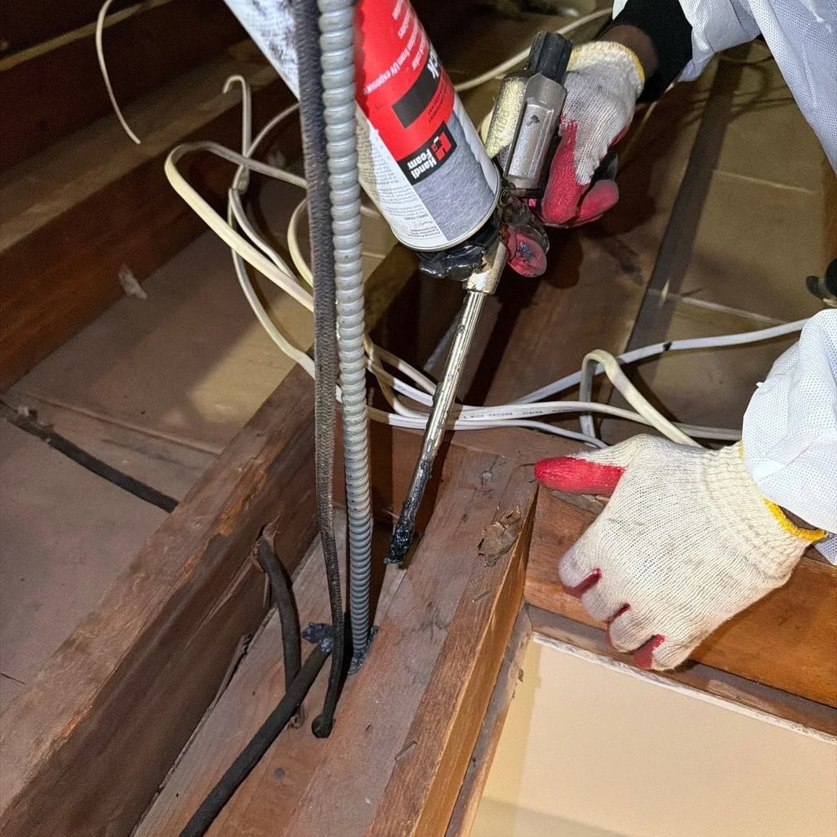 A gloved crew member applying foam sealant where wiring passes through attic framing